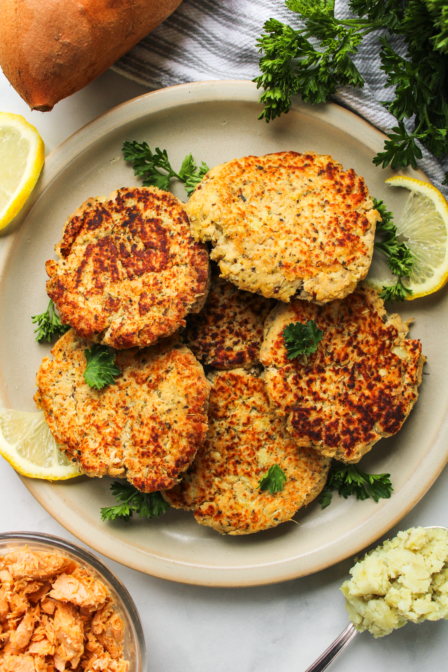 zoomed out shot of salmon sweet potato cakes on a grey ceramic plate topped with fresh parsley and ingredients surrounding the plate