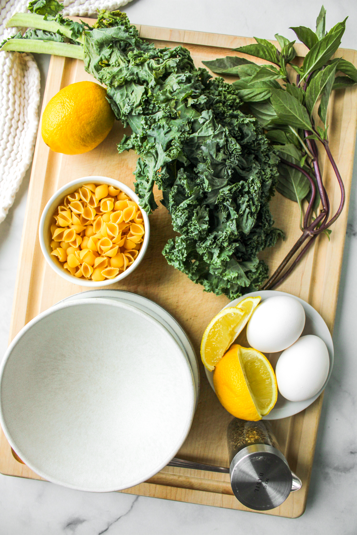 soup ingredients such as kale, basil, pasta shells, eggs, and lemon, displayed on a wooden board