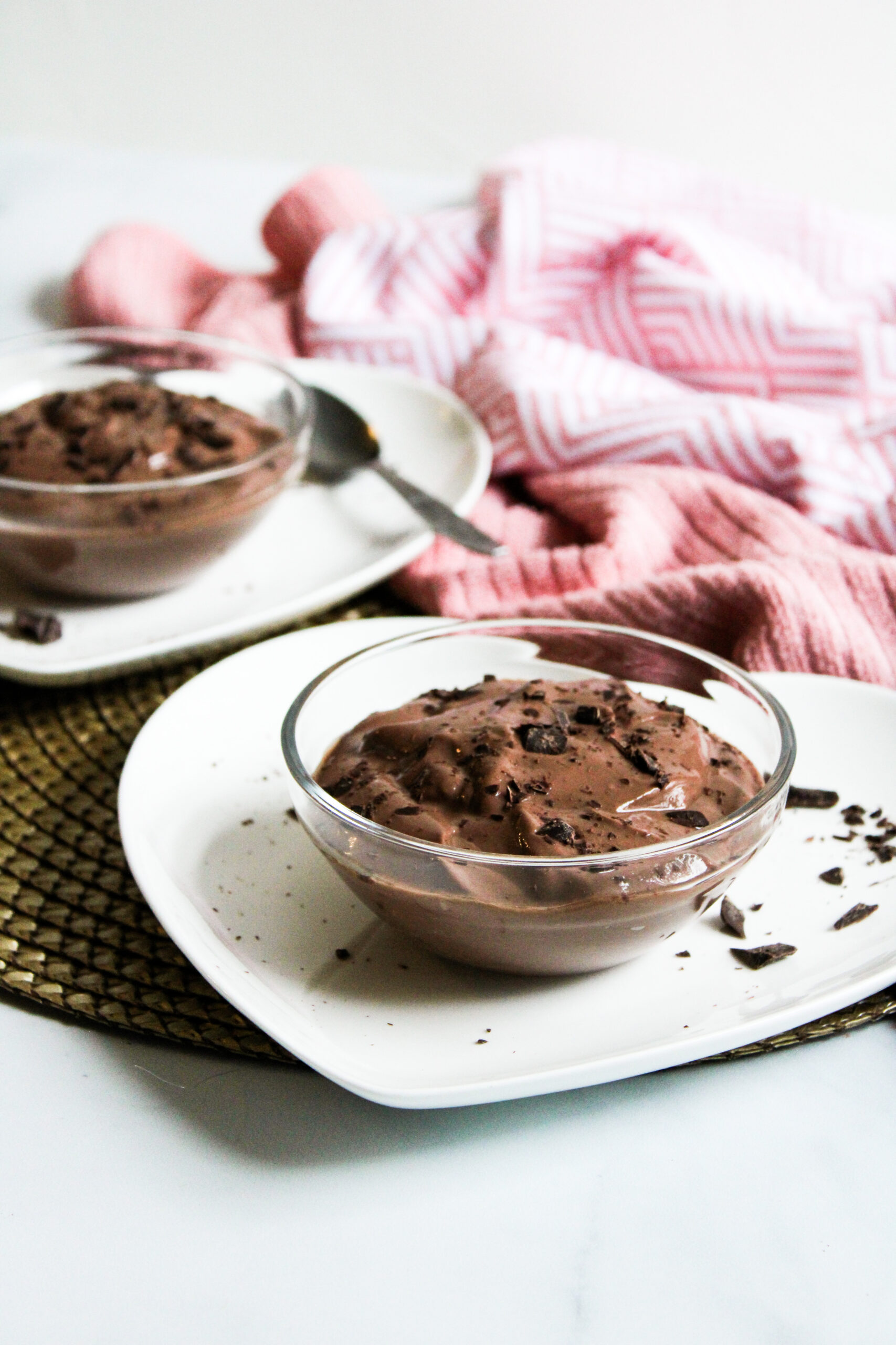 side profile of chocolate mousse in small glass bowls on top of white heart-shaped plates with a pink towel in the background