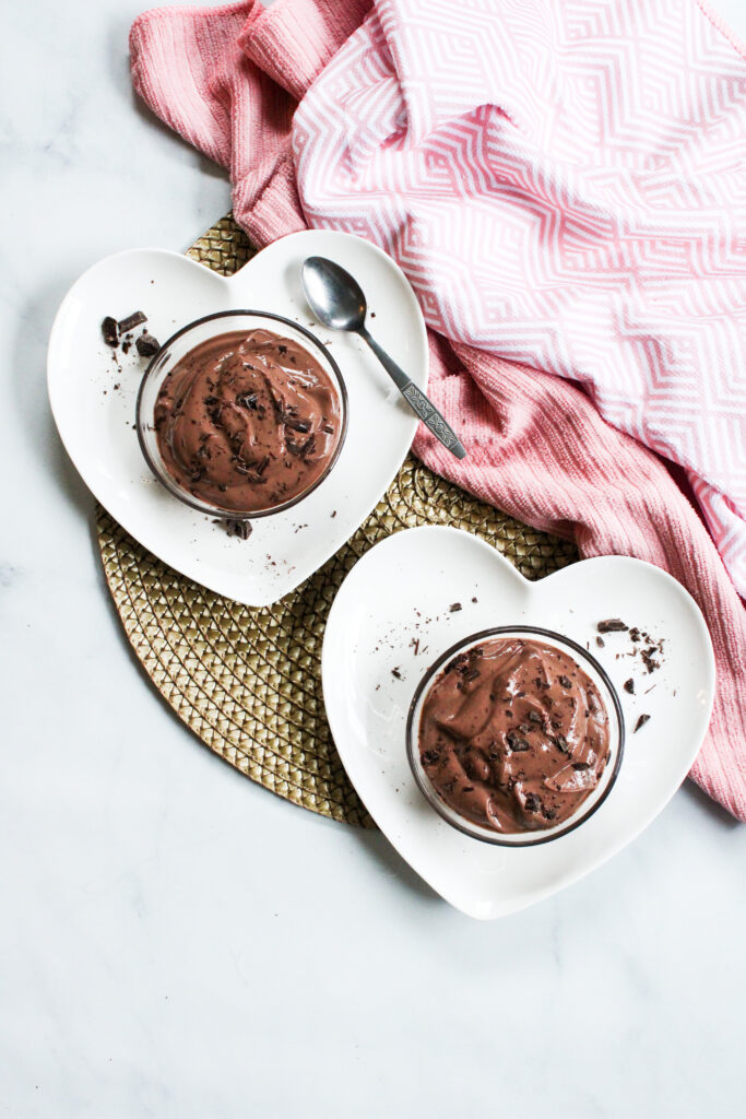 chocolate mousse in small glass bowls on top of white heart-shaped plates with a pink towel in the background