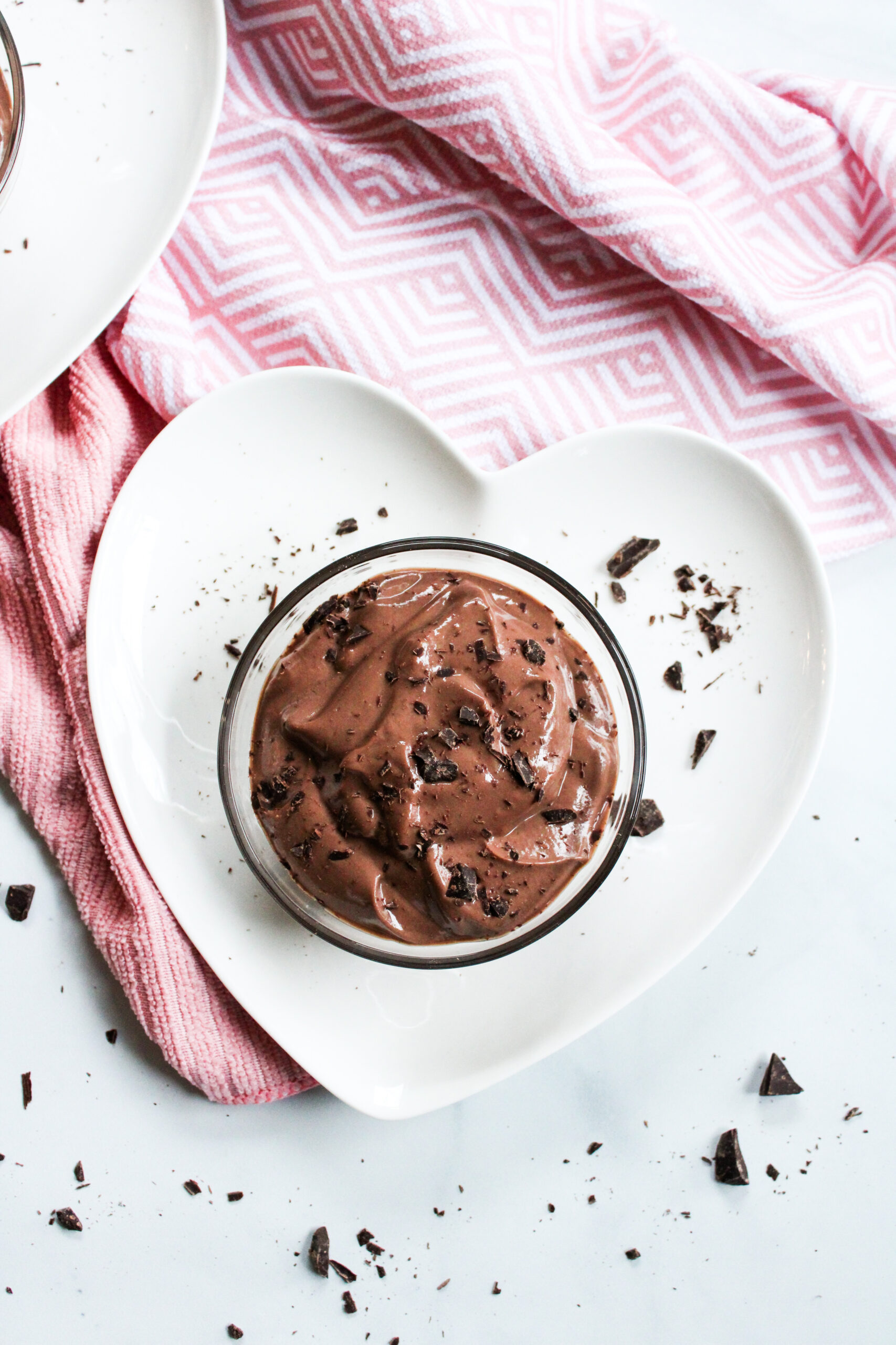 vegan chocolate mousse in a small glass bowl on top of white heart-shaped plates with a pink towel in the background