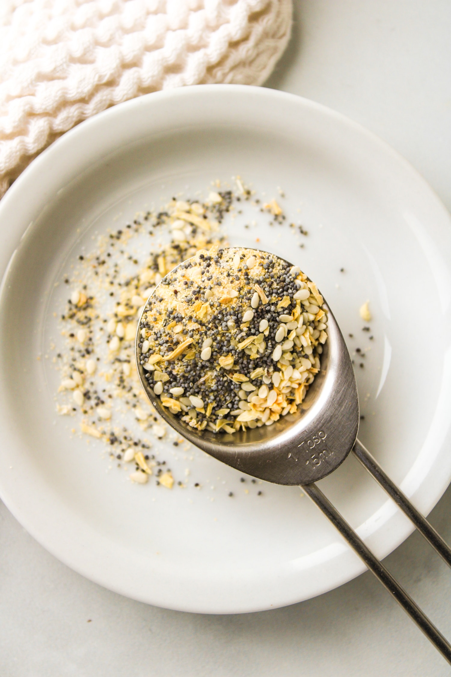 overhead shot of everything bagel seasoning in a stainless steel measuring spoon on top of a small white ceramic plate