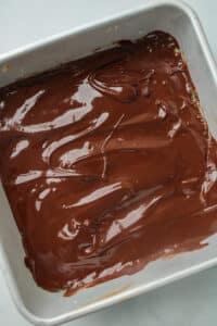 melted chocolate spread over nut bar mixture in a square stainless steel pan