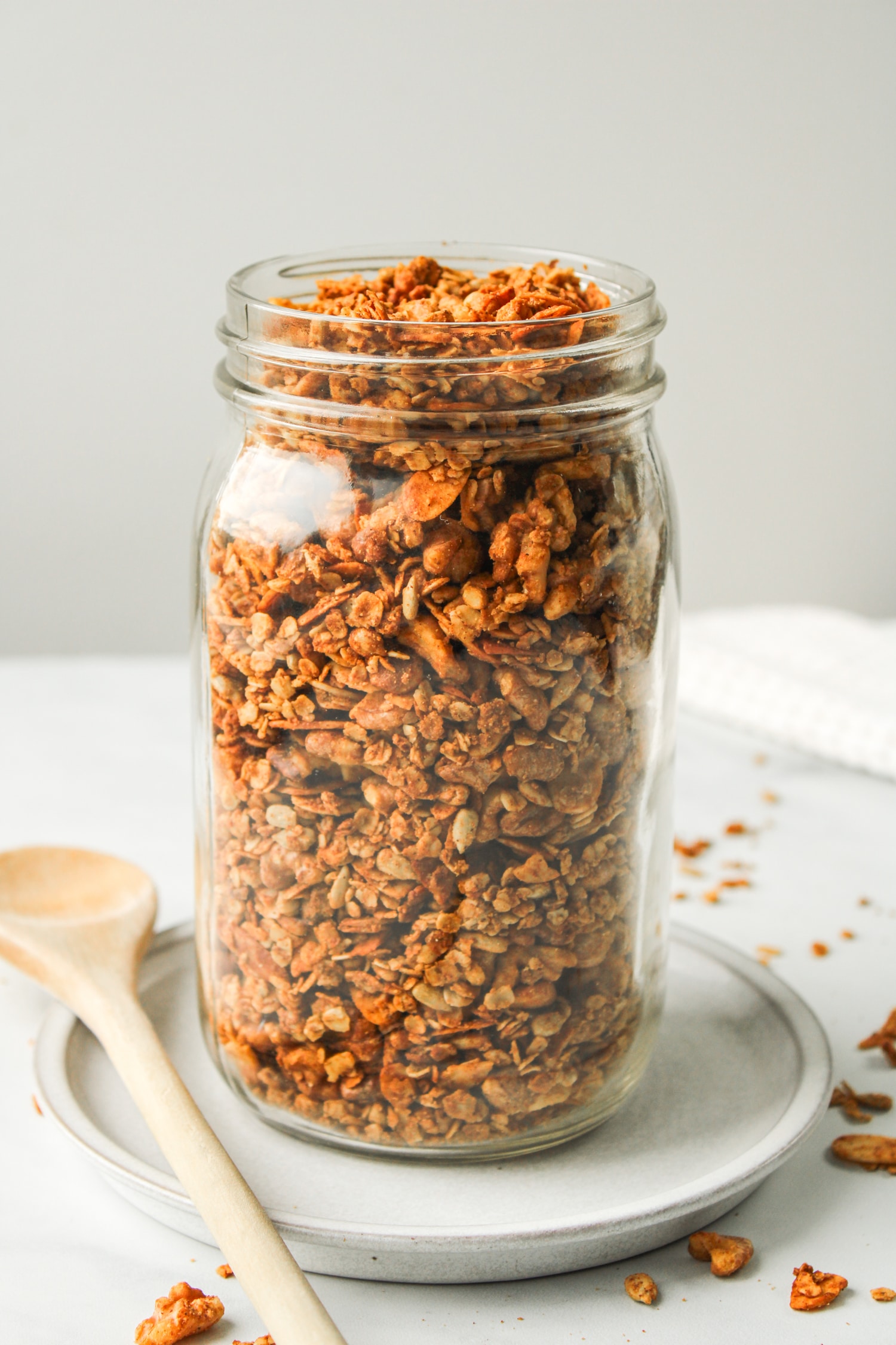 profile shot of air fryer granola in a large glass mason jar on top of a white ceramic dish with a wooden spoon beside it