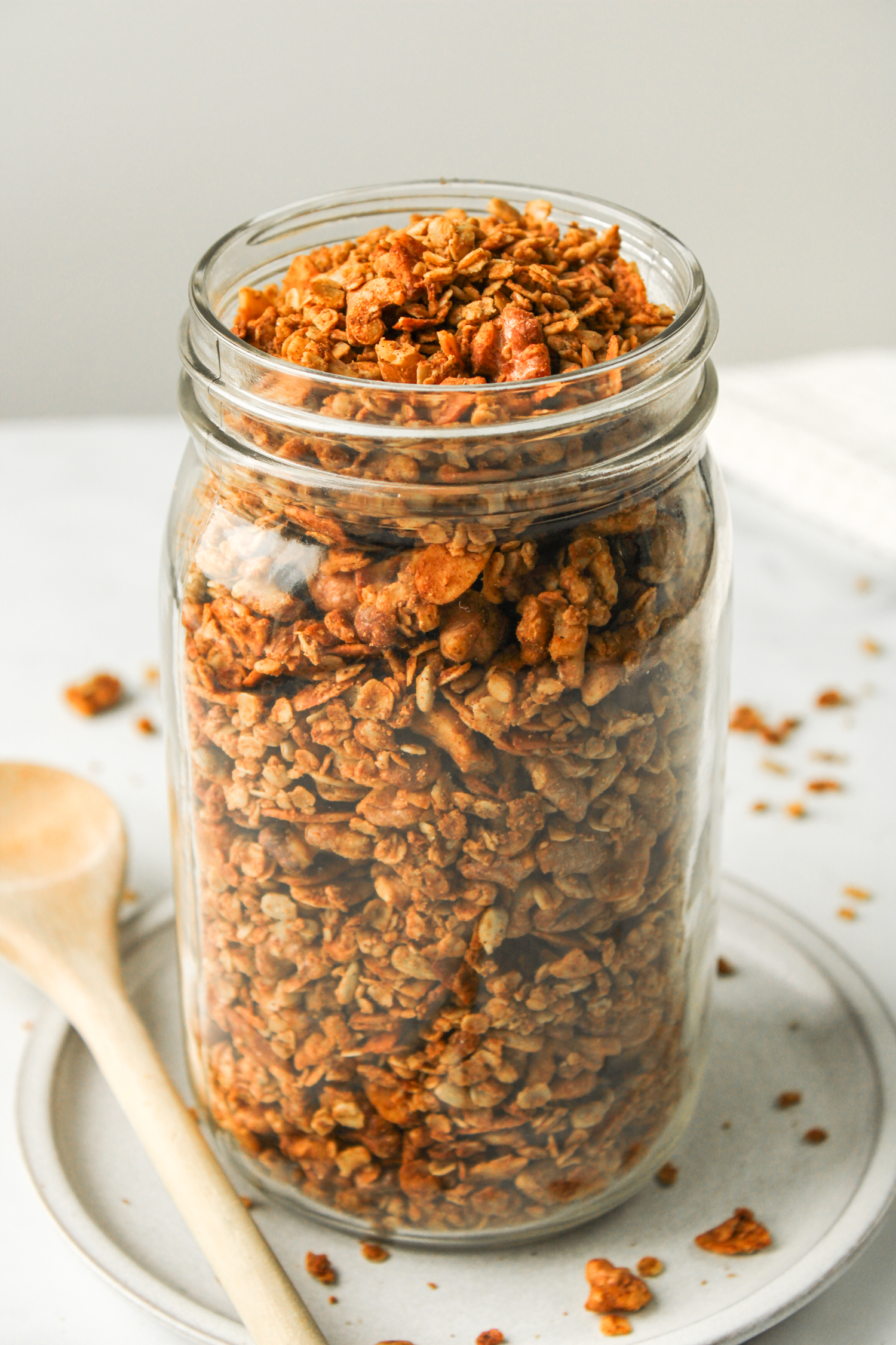 air fryer granola in a large glass mason jar on top of a white ceramic dish with a wooden spoon beside it