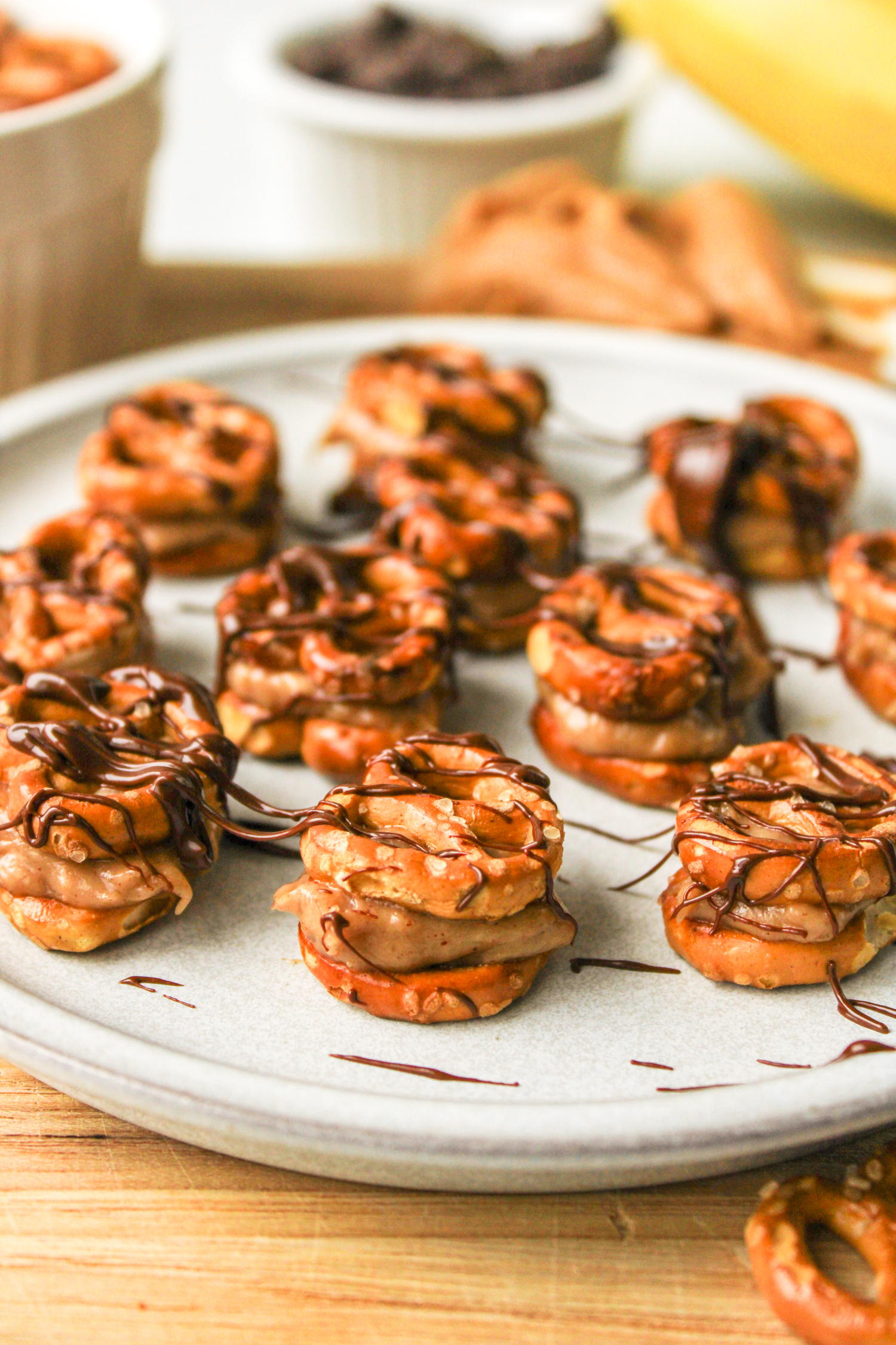 angled shot of pretzel peanut butter bites drizzled with chocolate on a grey ceramic plate, surrounded by recipe ingredients