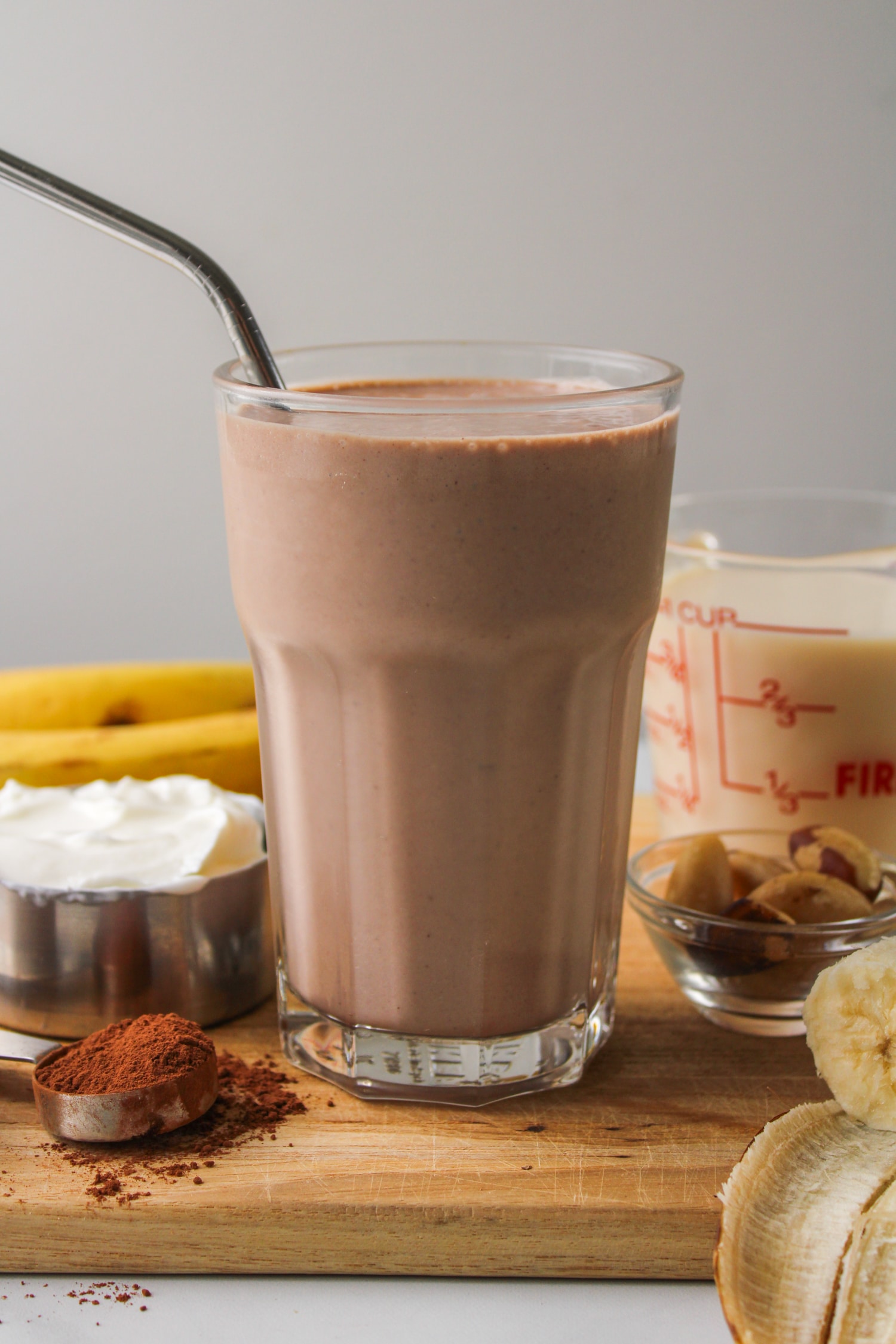 zoomed out photo of banana chocolate smoothie in a glass cup with a metal straw, surrounded by ingredients