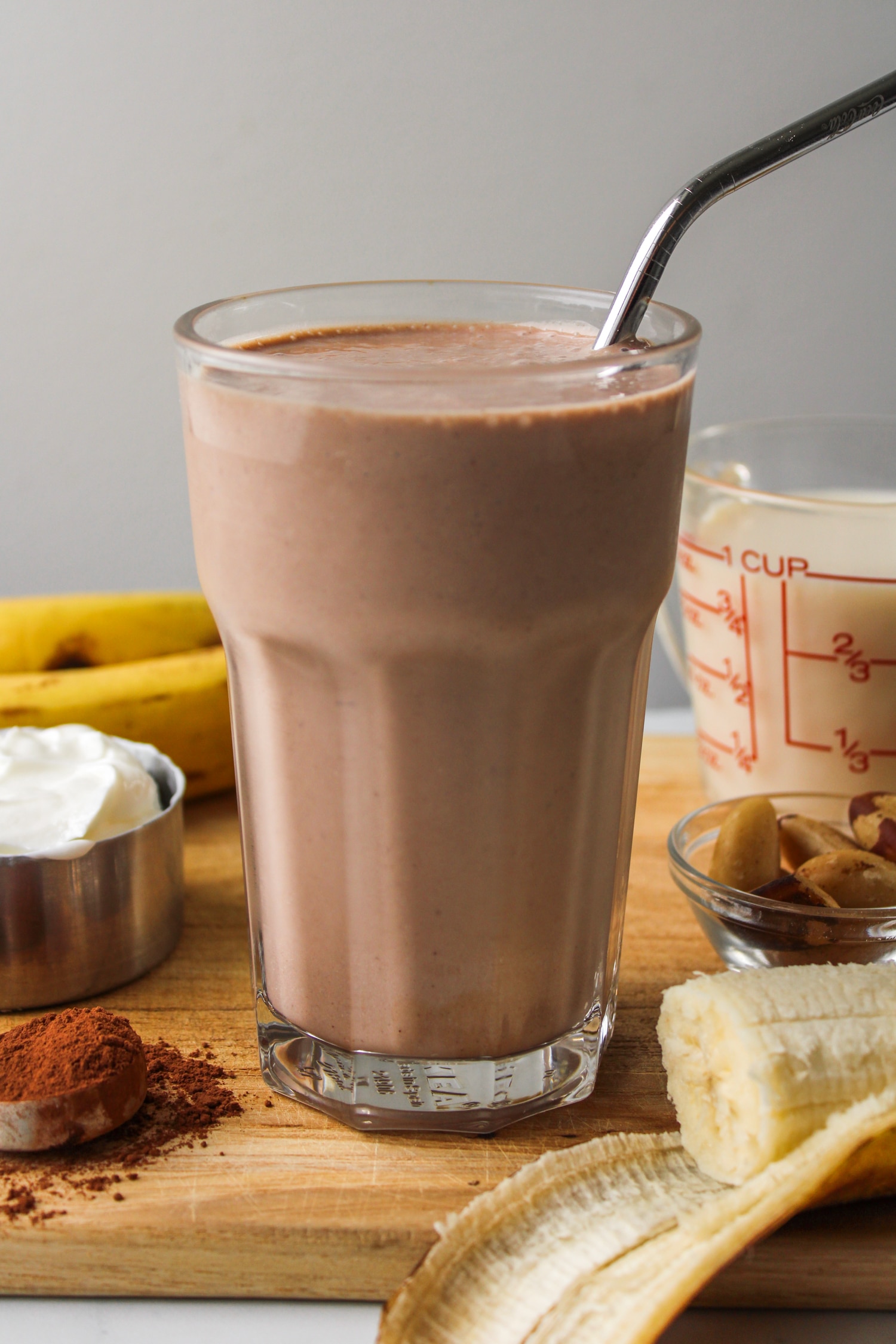 banana chocolate smoothie in a glass cup with a metal straw, surrounded by ingredients