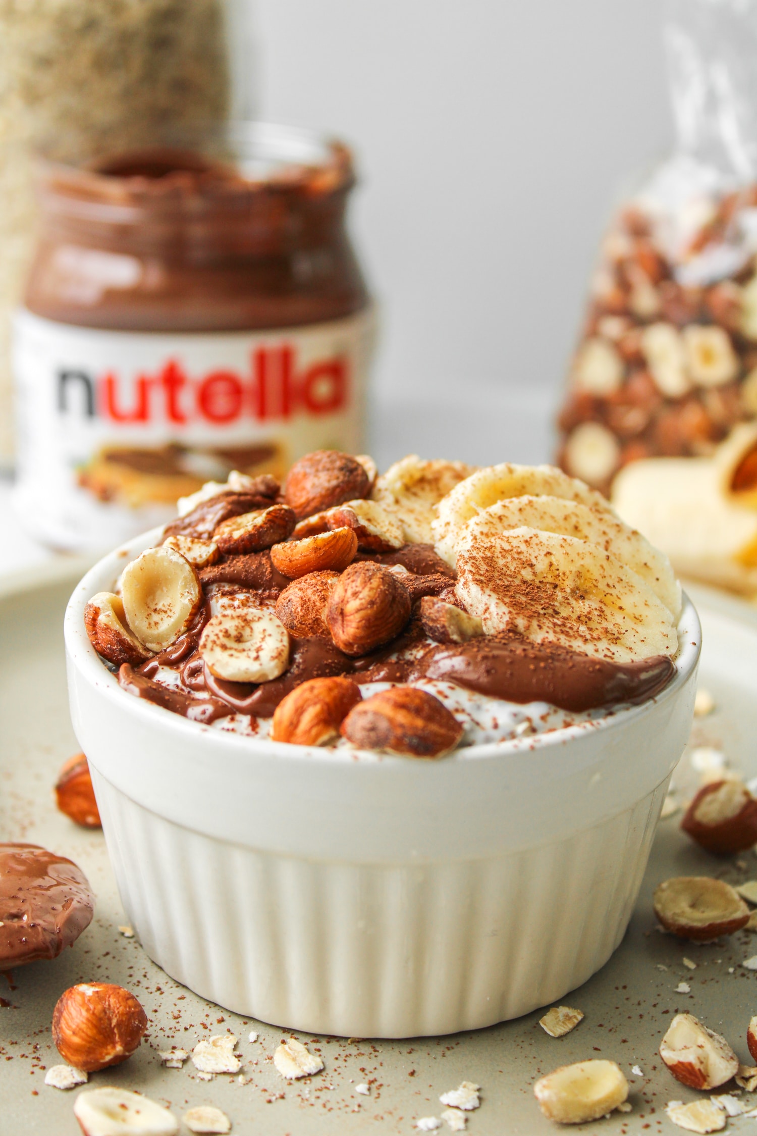 chocolate hazelnut overnight oats in a white ceramic dish topped with nutella, sliced banana, chopped hazelnuts and cocoa powder