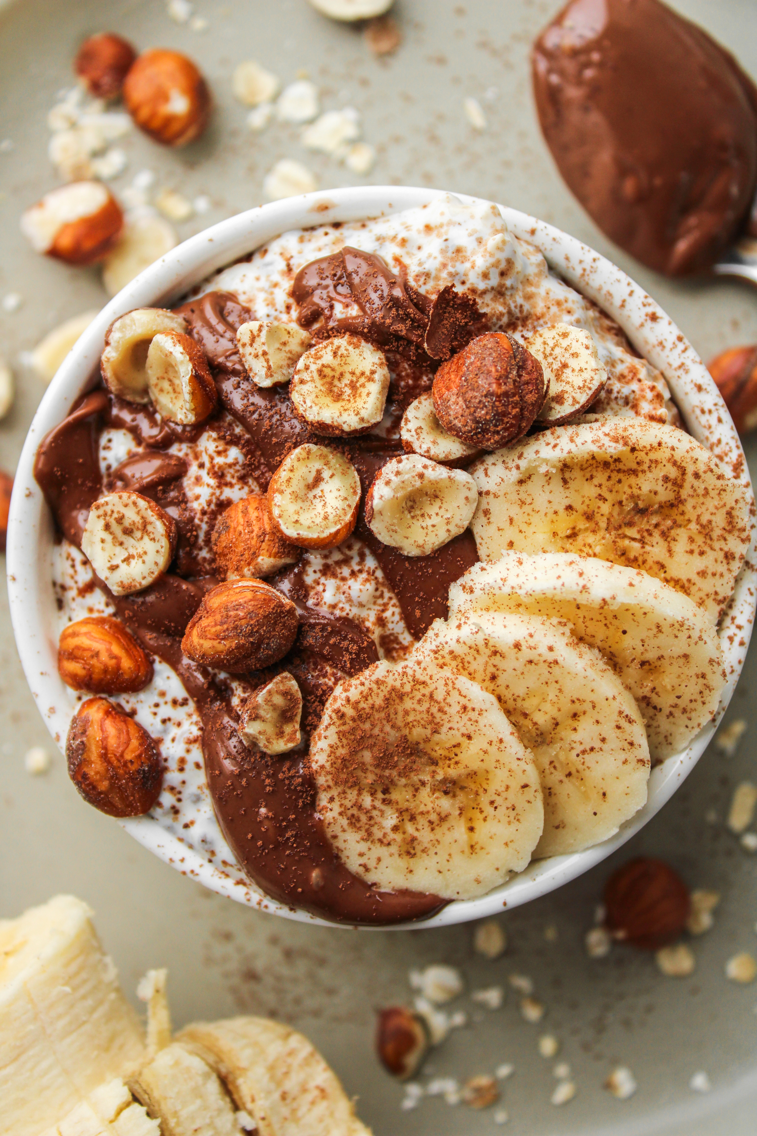 an overhead shot of nutella overnight oats in a white ceramic dish topped with nutella, sliced banana, chopped hazelnuts and cocoa powder