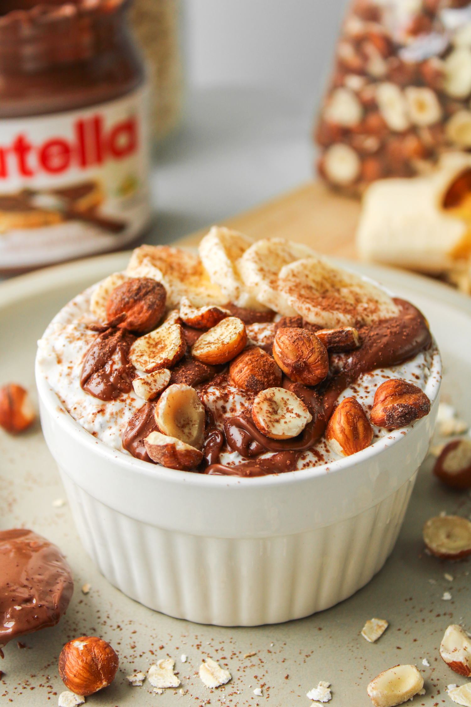 nutella overnight oats in a white ceramic dish topped with nutella, sliced banana, chopped hazelnuts and cocoa powder