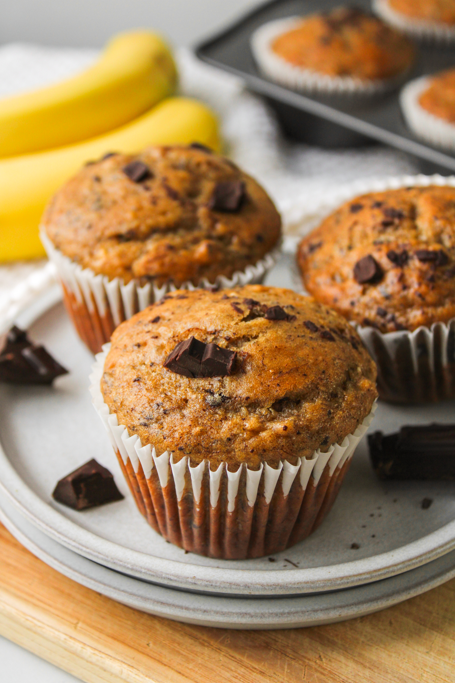 three banana chocolate chunk muffins on a grey ceramic plate with bananas and more muffins in the background