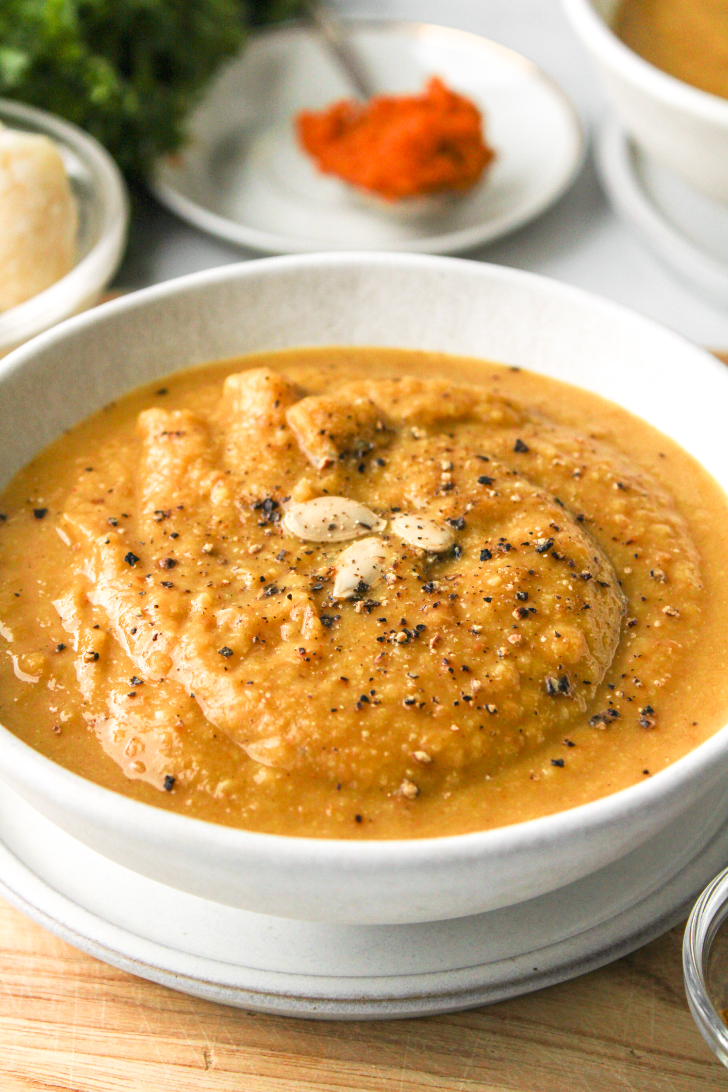 zoomed in side shot of cauliflower soup in white ceramic bowl on top of wooden board and ingredients in the background