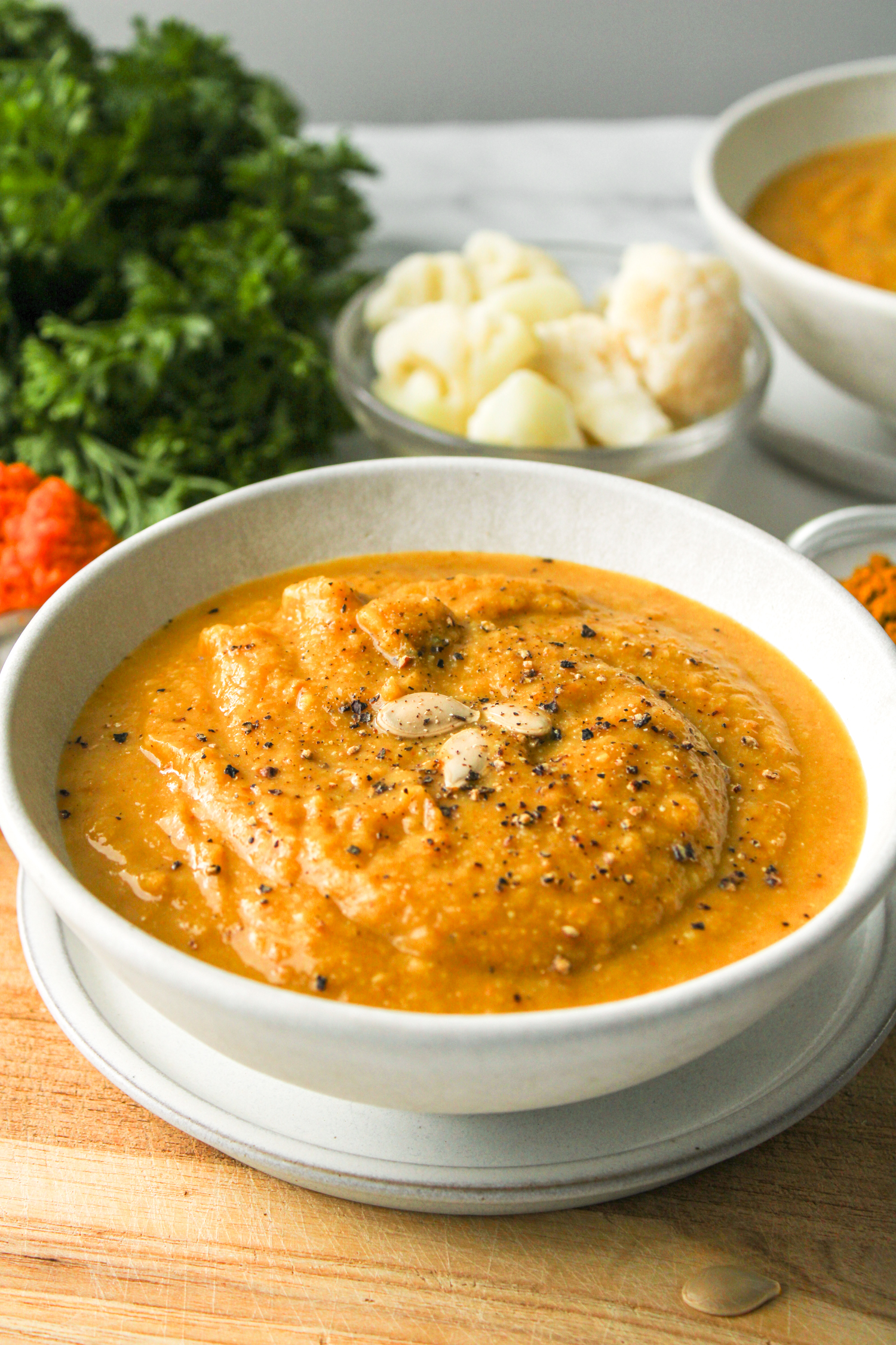 side shot of pumpkin soup in white ceramic bowl on top of wooden board and ingredients in the background