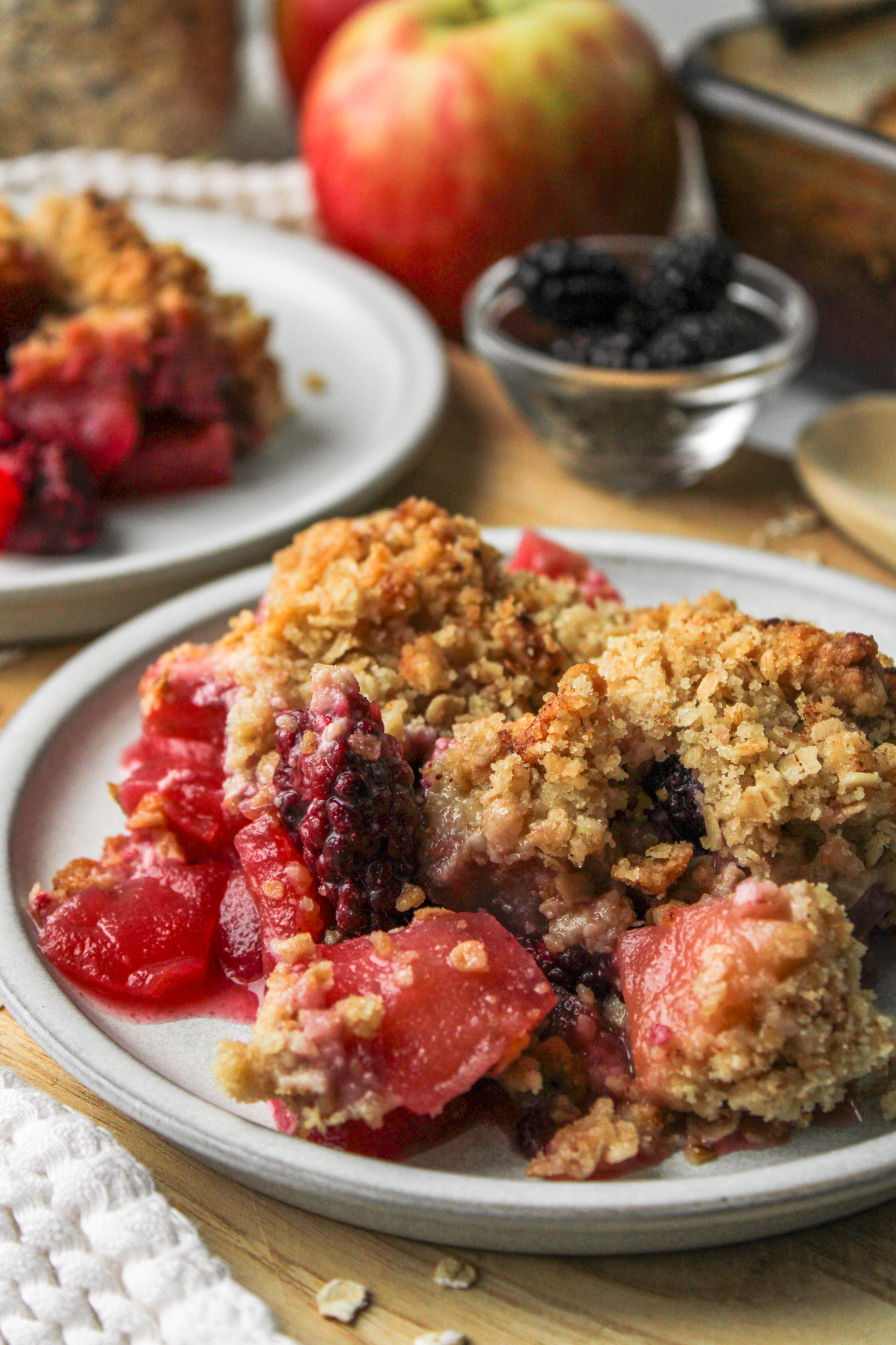 apple and blackberry crumble on a grey ceramic plate on top of a wooden cutting board with crumble and ingredients in the background