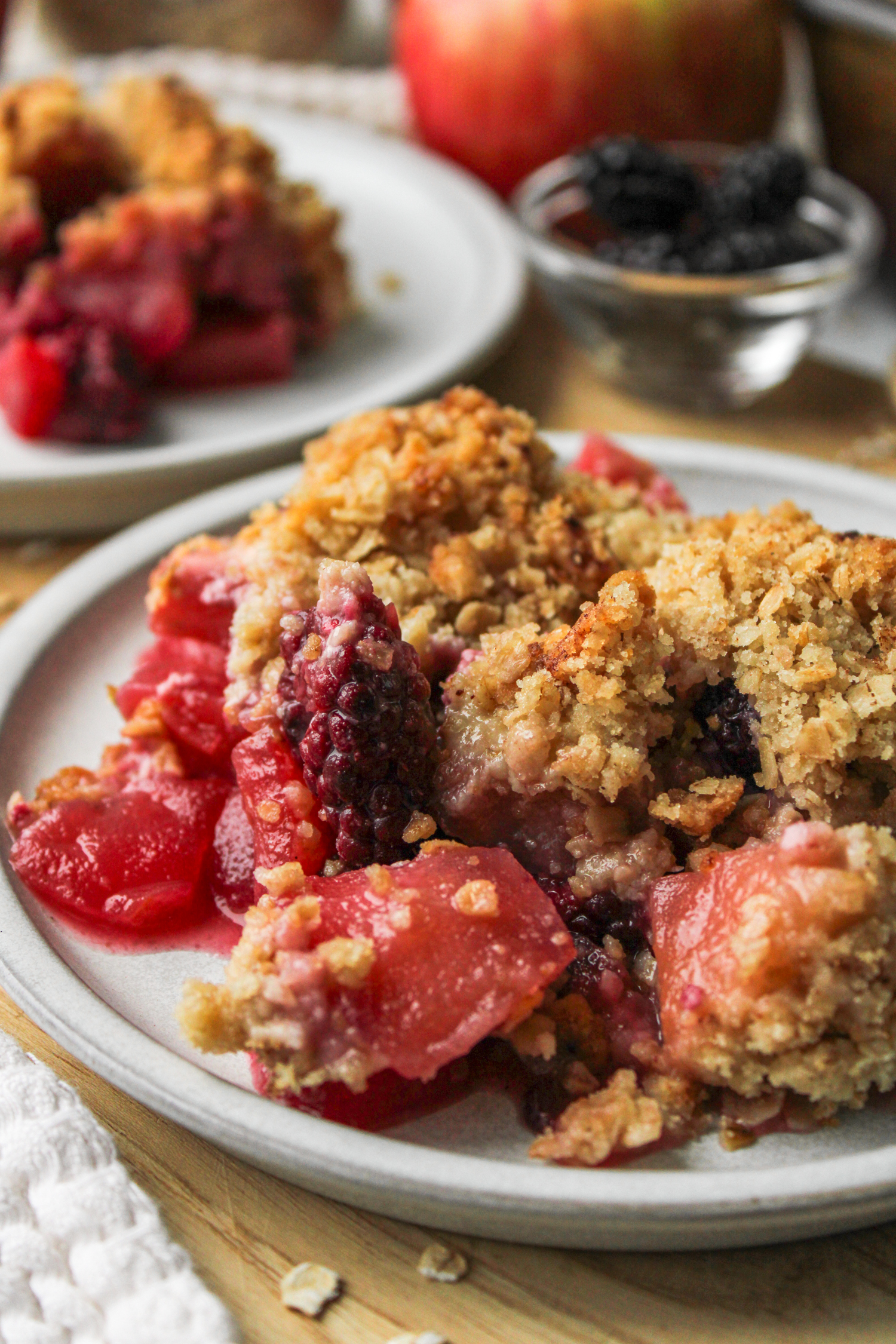 close up photo of apple blackberry crumble on a grey ceramic plate on top of a wooden cutting board with crumble and ingredients in the background