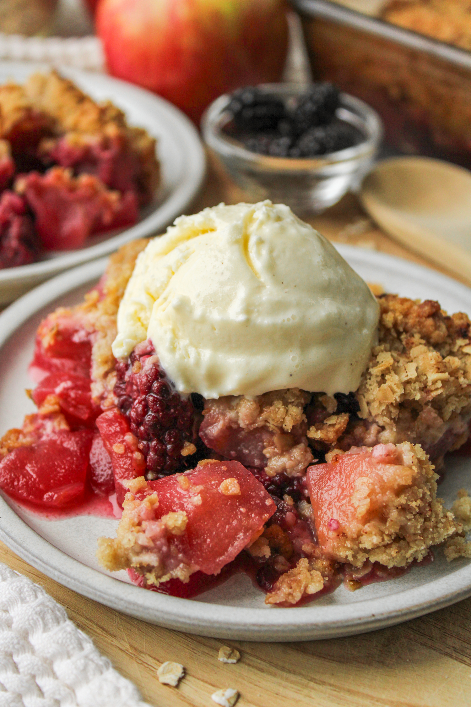 apple and blackberry crumble with ice cream on top on a grey ceramic plate on top of a wooden cutting board with crumble and ingredients in the background