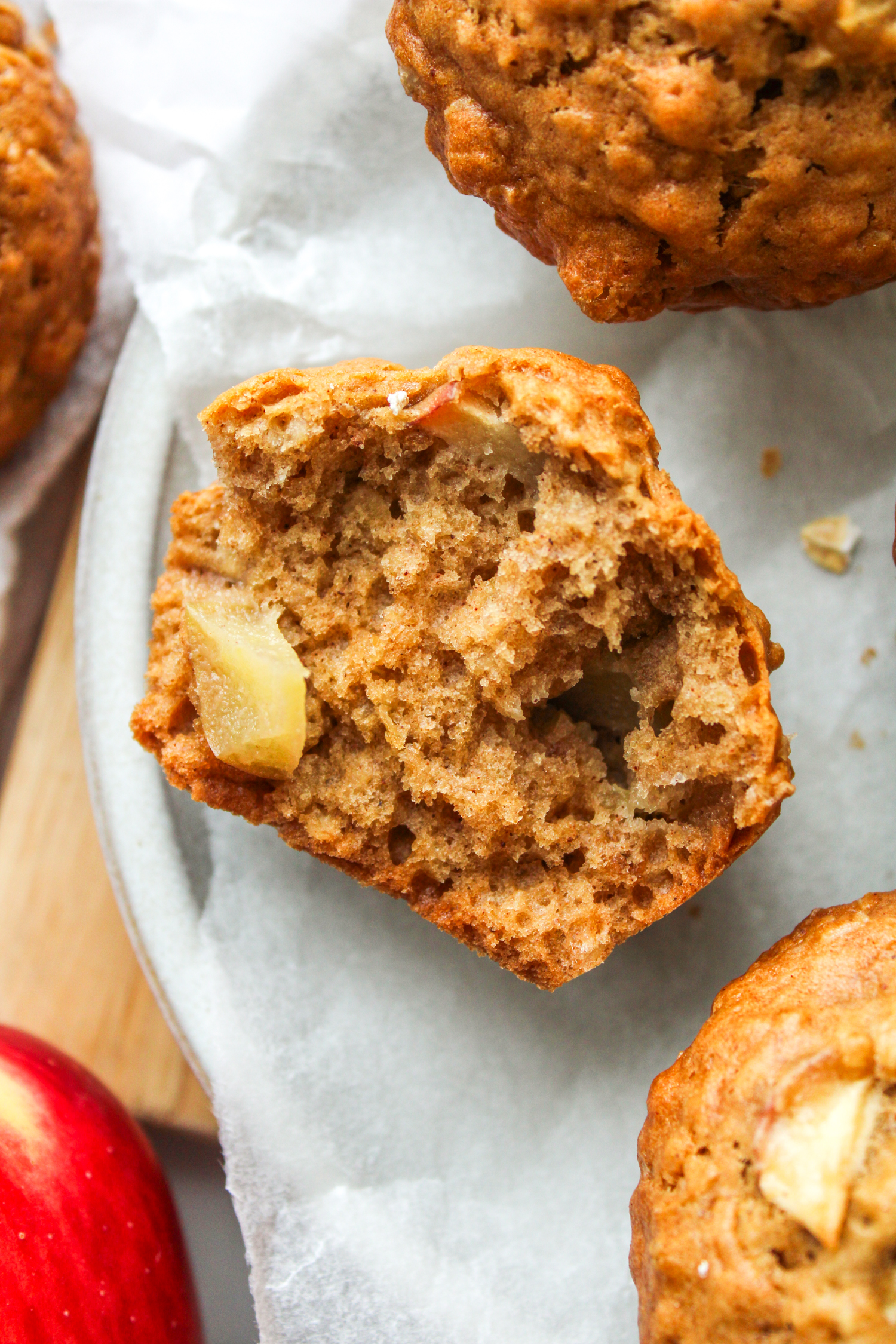 overhead shot of half of an apple oatmeal muffin face up on top of parchment paper on a white ceramic plate with whole muffins around it