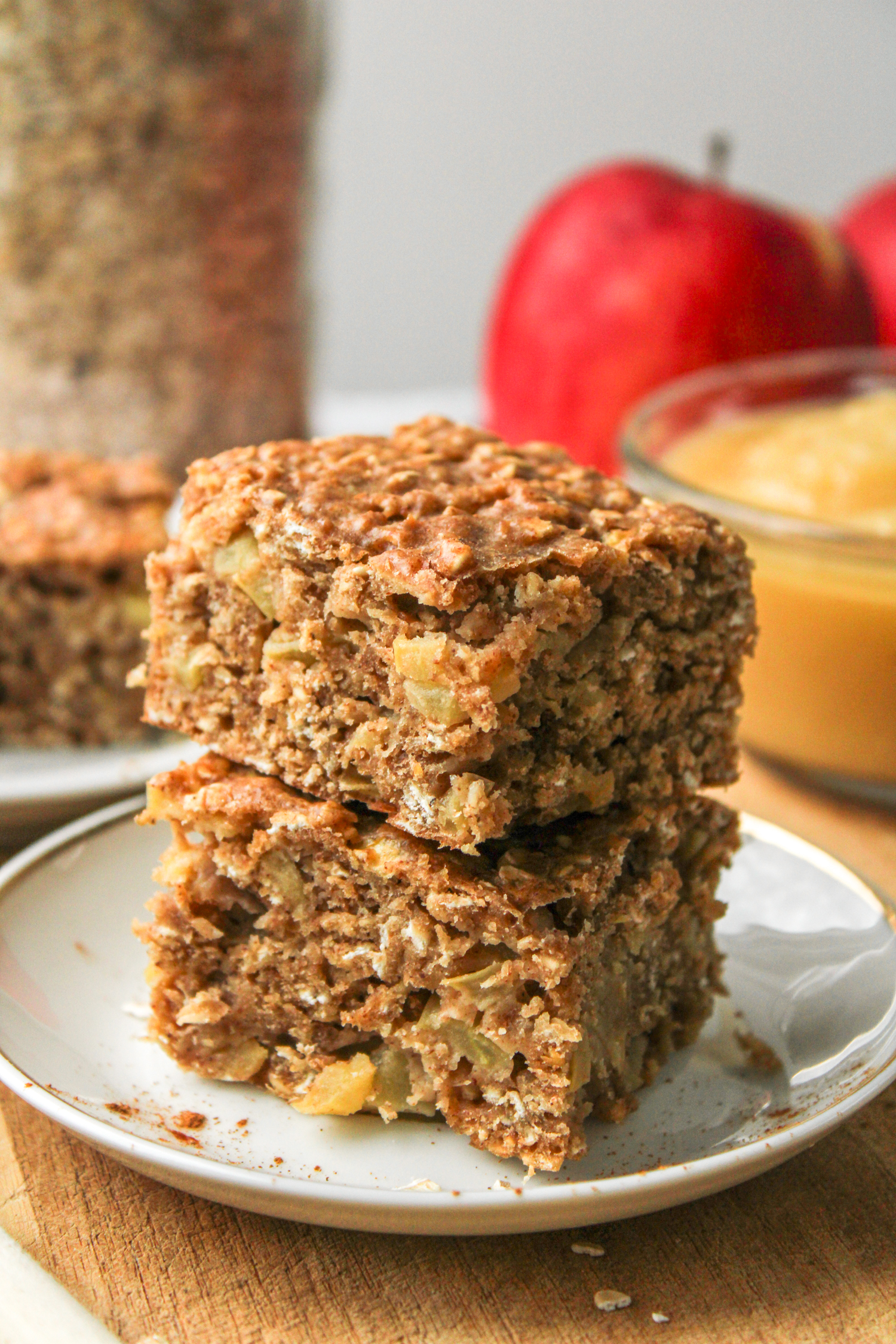 zoomed in photo of two applesauce oatmeal bars stacked on top of each other on a small white ceramic plate with ingredients in the background