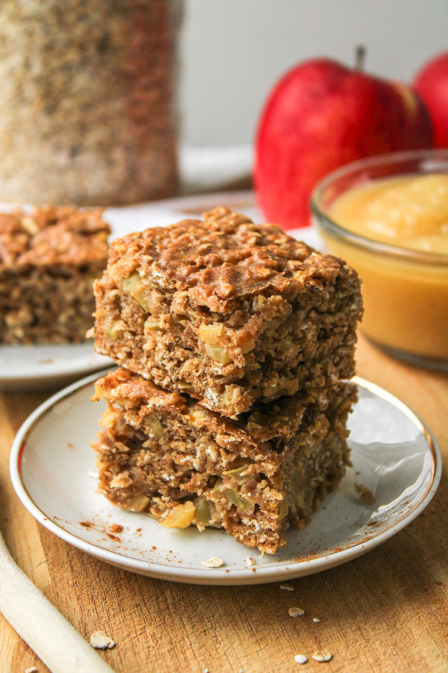 two applesauce oatmeal bars stacked on top of each other on a small white ceramic plate with ingredients in the background