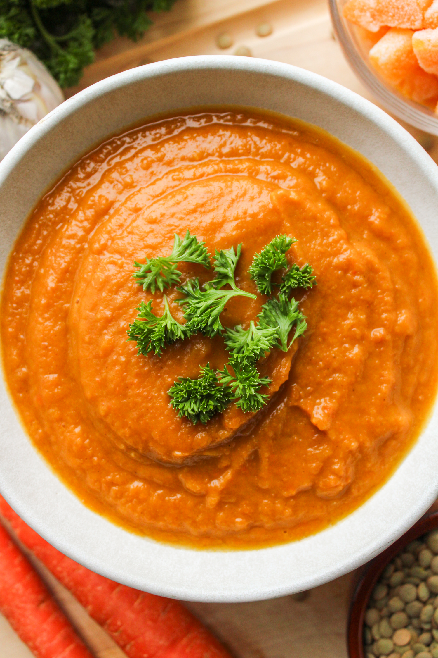 close up shot of butternut squash and carrot soup topped with green parsley, in a white ceramic bowl on top of a wooden cutting board