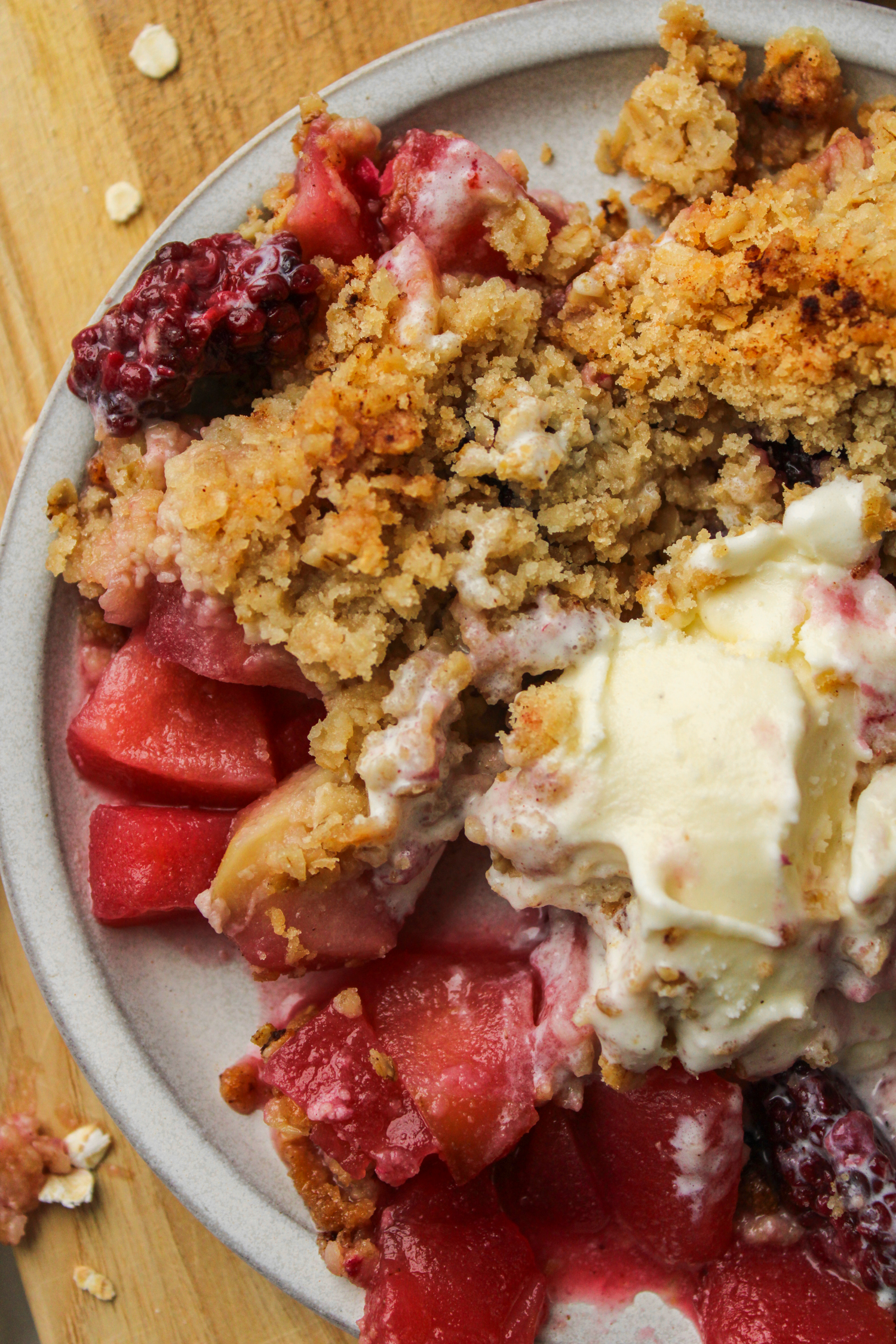 overhead shot of apple crumble with ice cream on top on a grey ceramic plate on top of a wooden cutting board