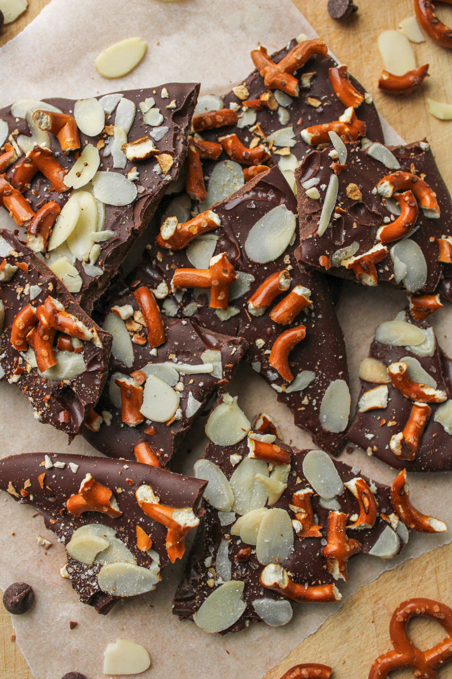 pretzel almond bark pieces on top of a wooden cutting board with pieces of pretzels, almonds and chocolate around the bark