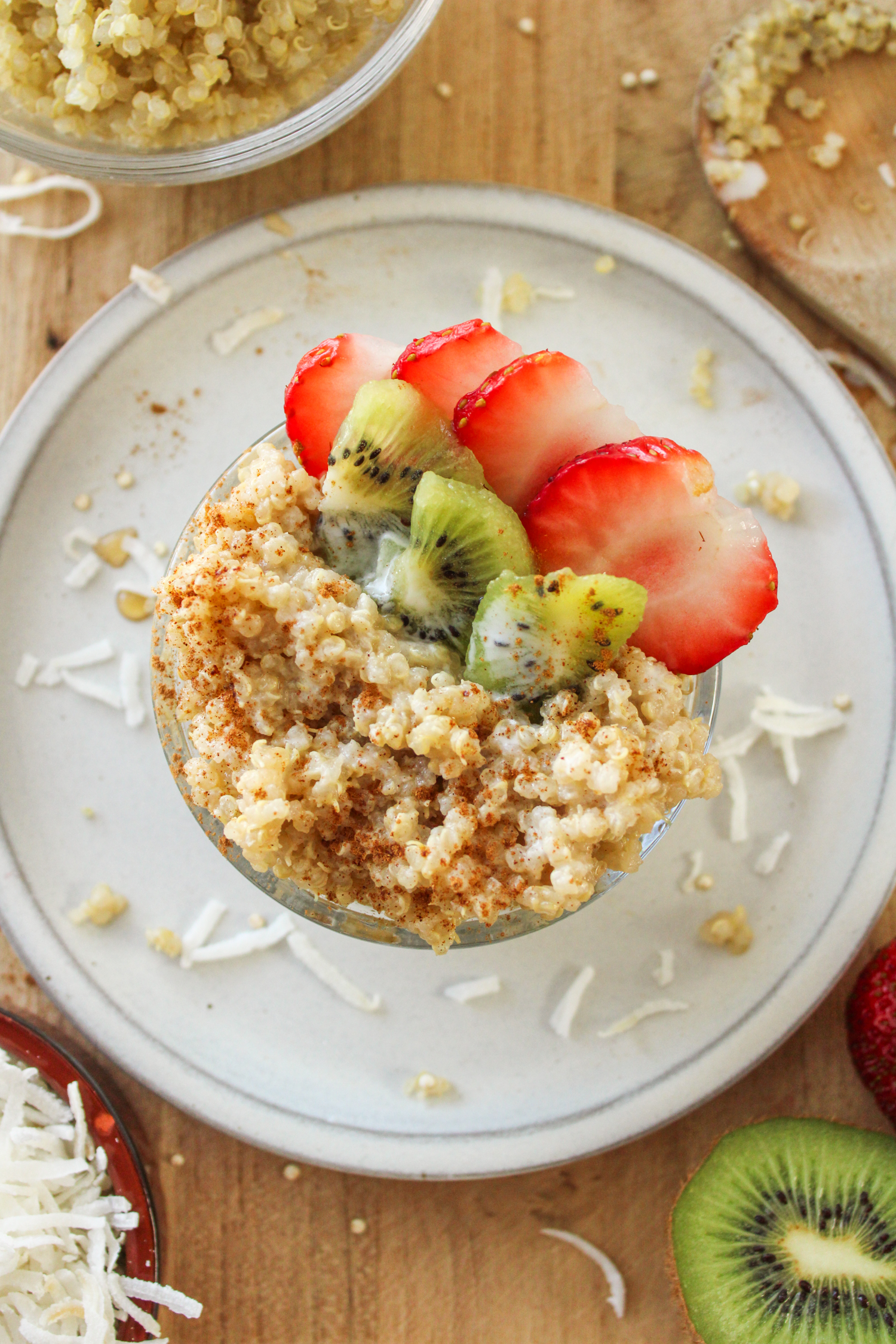 overhead shot of quinoa coconut porridge in a small glass bowl topped with kiwi and strawberries on a grey ceramic plate