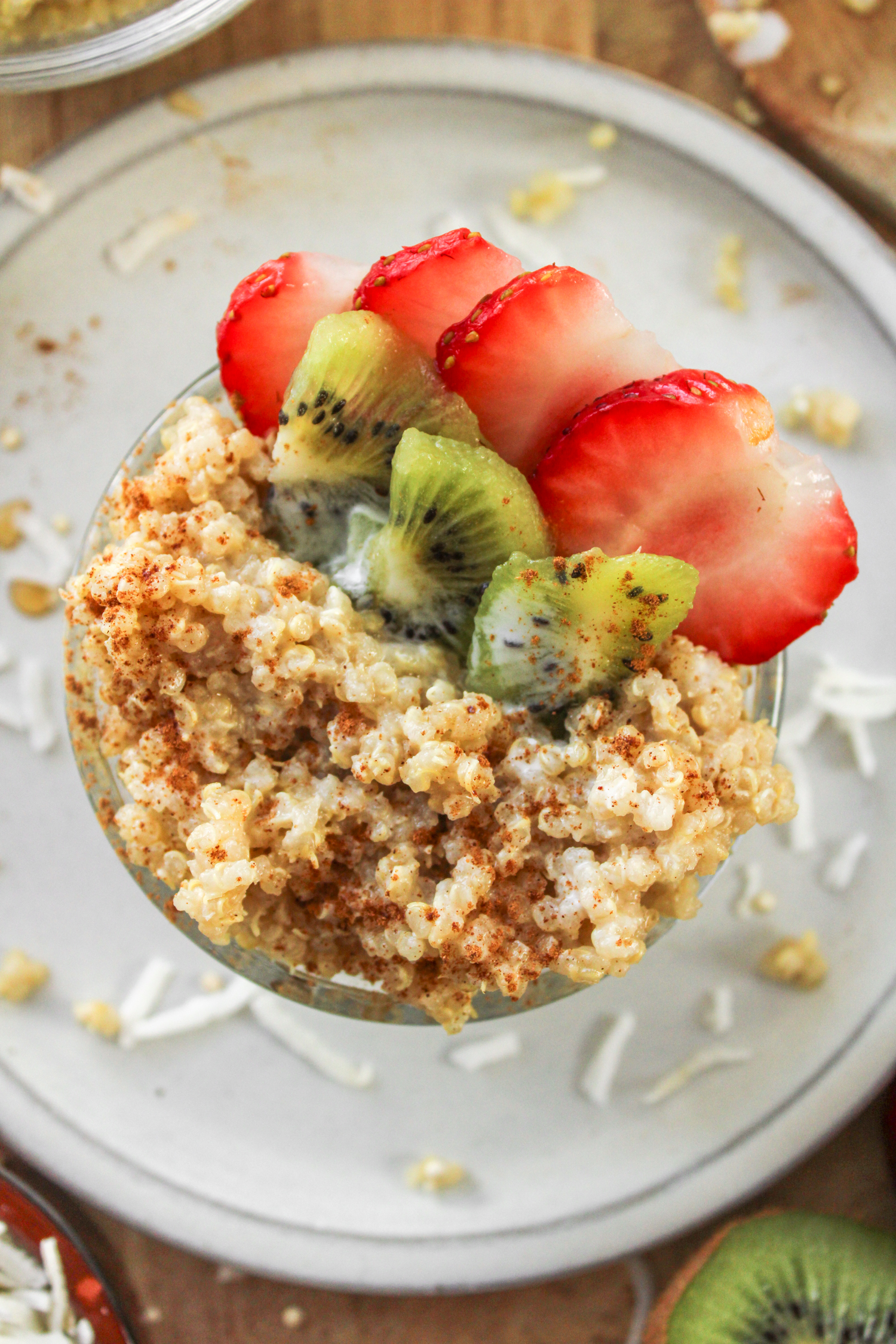 close up shot of overhead shot of coconut quinoa porridge in a small glass bowl topped with kiwi and strawberries on a grey ceramic plate
