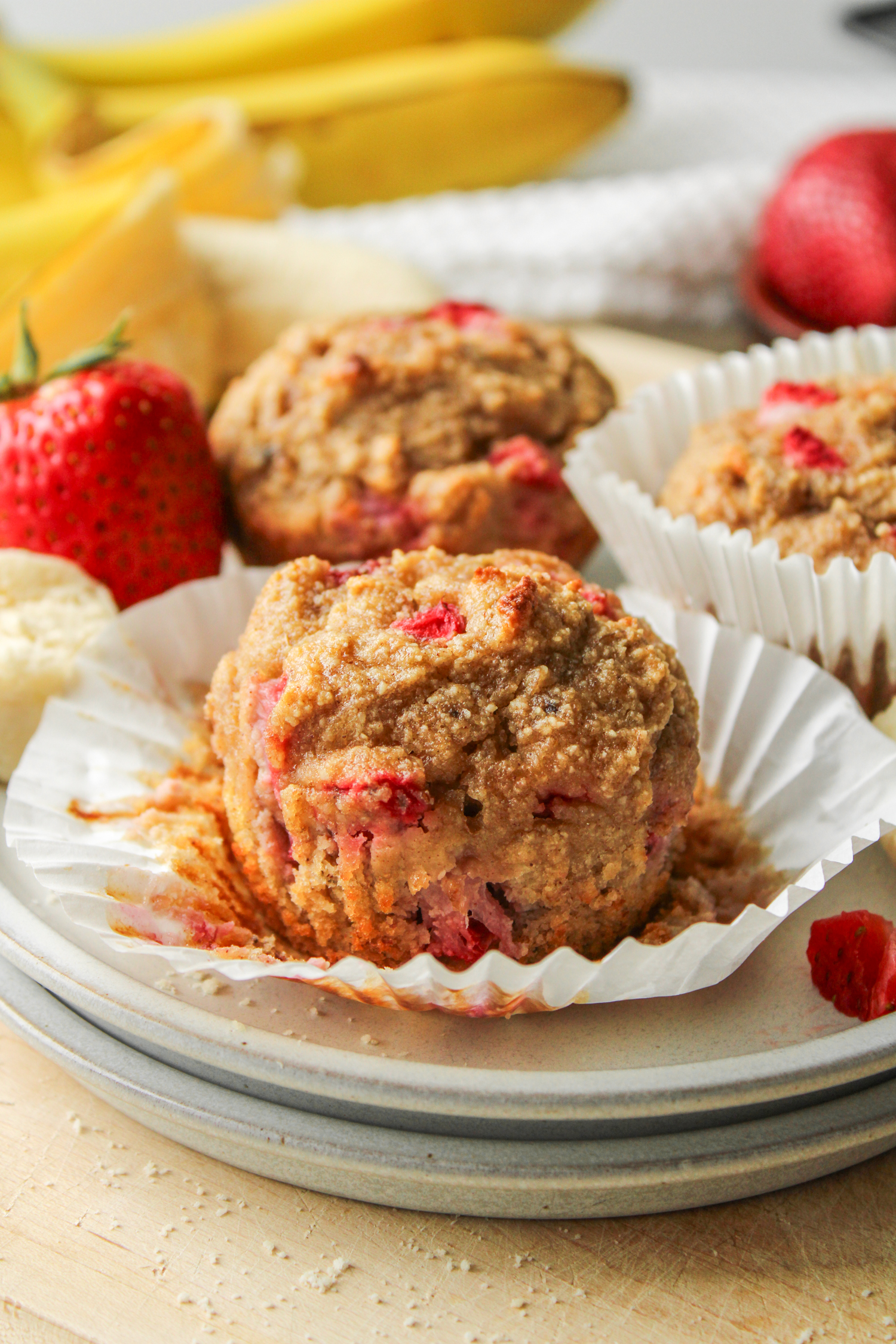 strawberry banana and almond flour muffins on a paper muffin liner on top of two grey ceramic plates with muffins in the background